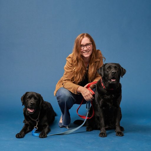Elyse Manterfield squatting down between two black labrador retrievers